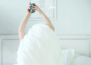 A person holding a coffee mug while relaxing in bed, covered with a white sheet.