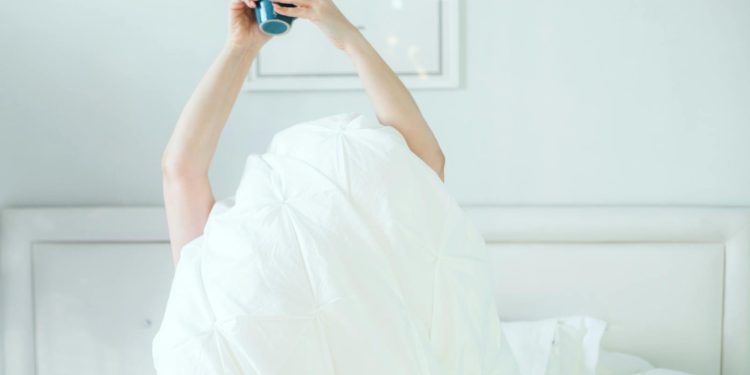 A person holding a coffee mug while relaxing in bed, covered with a white sheet.