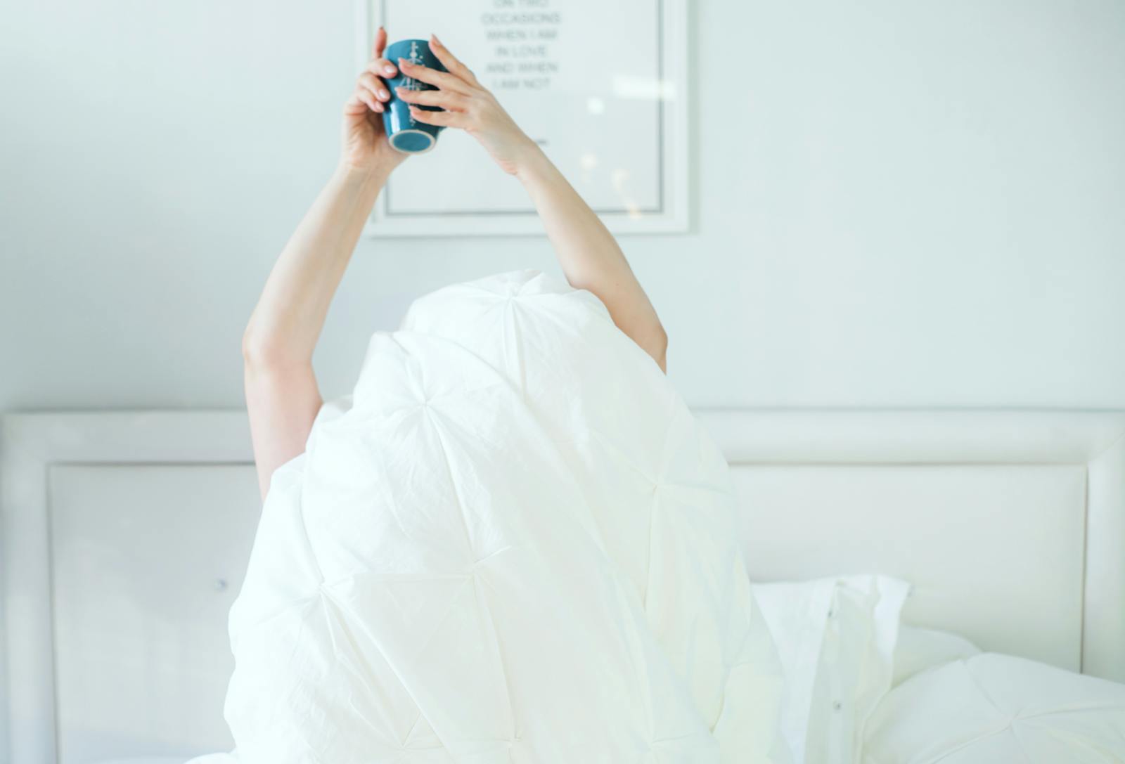 A person holding a coffee mug while relaxing in bed, covered with a white sheet.