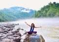 A smiling woman sits on a rock by a misty river, surrounded by lush mountains, enjoying nature.