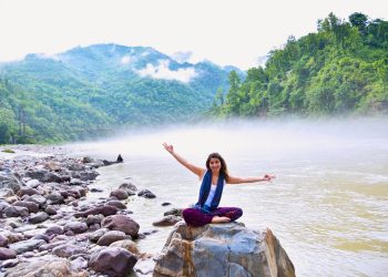 A smiling woman sits on a rock by a misty river, surrounded by lush mountains, enjoying nature.