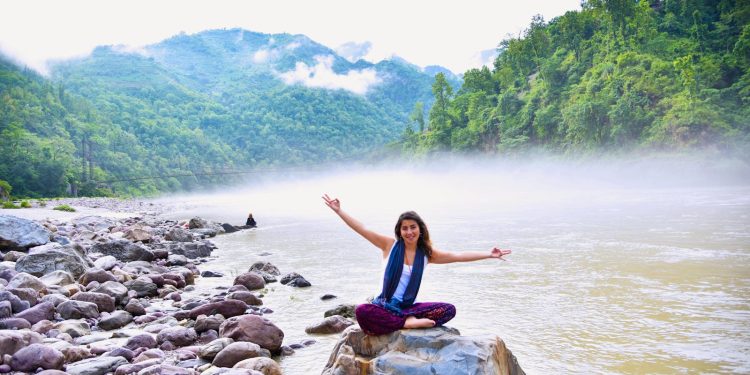 A smiling woman sits on a rock by a misty river, surrounded by lush mountains, enjoying nature.