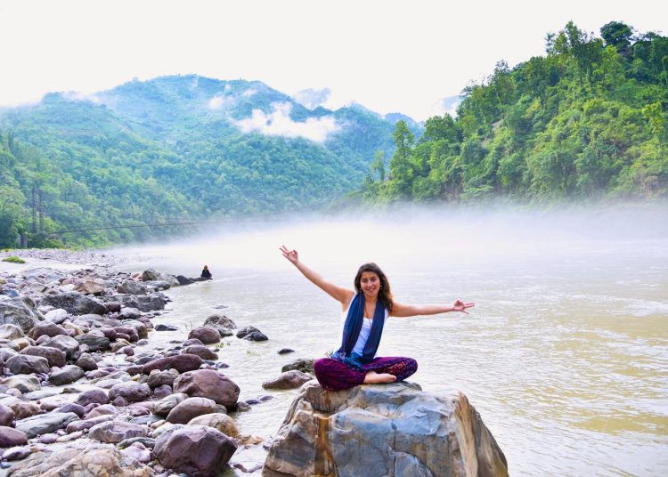 A smiling woman sits on a rock by a misty river, surrounded by lush mountains, enjoying nature.