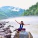 A smiling woman sits on a rock by a misty river, surrounded by lush mountains, enjoying nature.