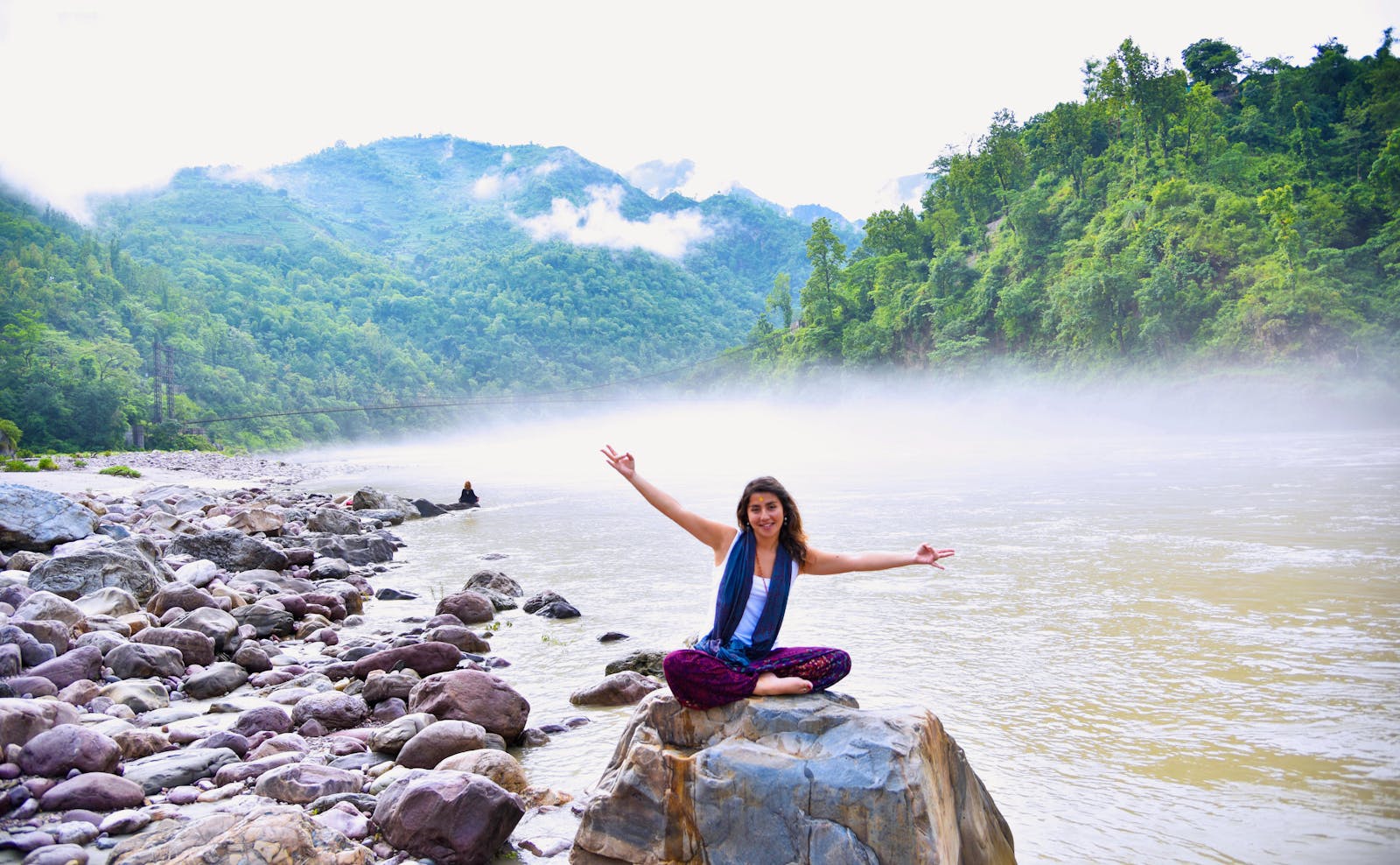 A smiling woman sits on a rock by a misty river, surrounded by lush mountains, enjoying nature.