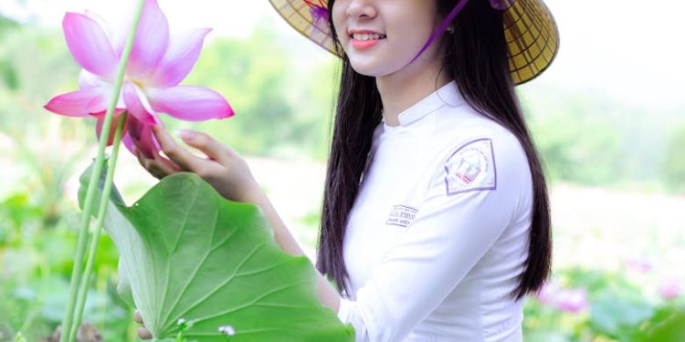 A young woman in traditional attire admires lotus flowers in a lush green field.