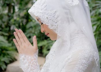 A serene moment of a Muslim woman in a bridal hijab, praying outdoors amidst lush greenery.