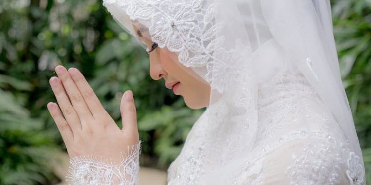 A serene moment of a Muslim woman in a bridal hijab, praying outdoors amidst lush greenery.