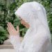 A serene moment of a Muslim woman in a bridal hijab, praying outdoors amidst lush greenery.