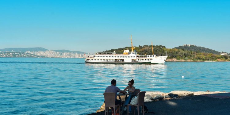 A couple enjoys the coastal view of İstanbul with a ferry in the background on a sunny day.