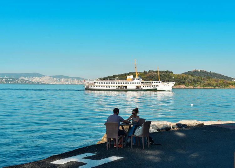 A couple enjoys the coastal view of İstanbul with a ferry in the background on a sunny day.