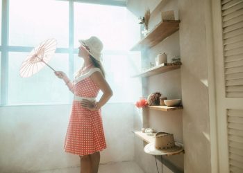 A woman in a vintage polka-dot dress holds a parasol by a sunlit window indoors.