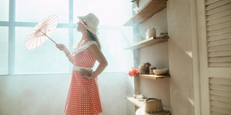 A woman in a vintage polka-dot dress holds a parasol by a sunlit window indoors.