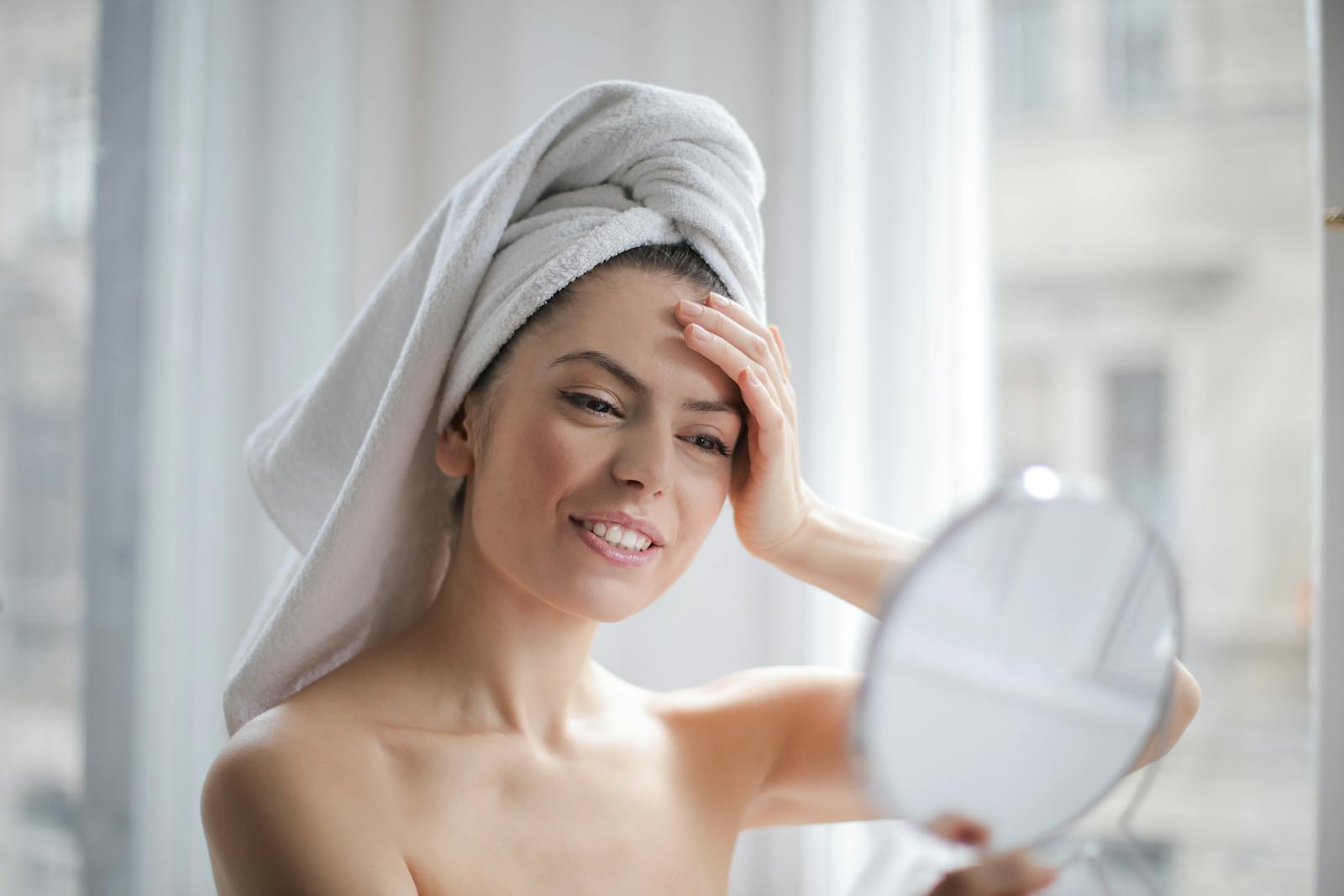 A woman with a towel on her head looking in a handheld mirror and smiling.