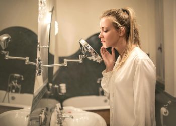 Side view of blond woman in white blouse standing in bathroom looking closely at face skin in small mirror