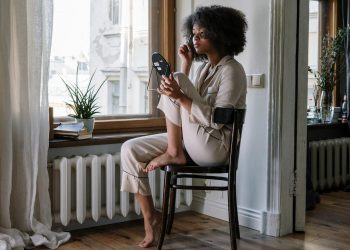 Woman in pajamas applying skincare with mirror, enjoying a cozy morning by the window.