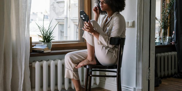 Woman in pajamas applying skincare with mirror, enjoying a cozy morning by the window.