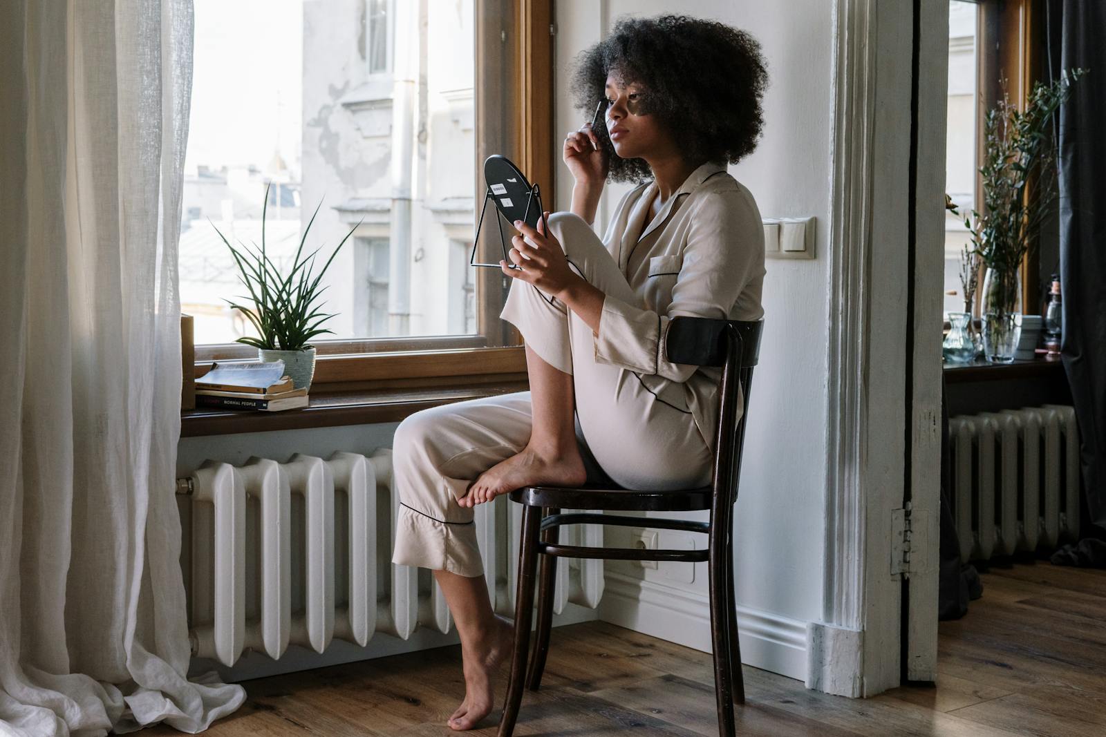 Woman in pajamas applying skincare with mirror, enjoying a cozy morning by the window.