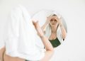 Woman with towel on head applies skincare, reflects self-care routine in mirror.