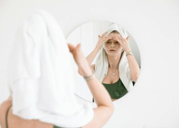 Woman with towel on head applies skincare, reflects self-care routine in mirror.
