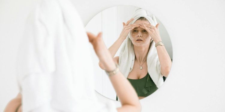 Woman with towel on head applies skincare, reflects self-care routine in mirror.