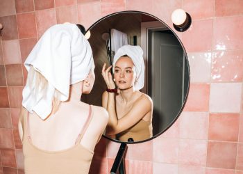 A young woman with a towel in a pink bathroom, reflecting in round mirror as she performs skincare.