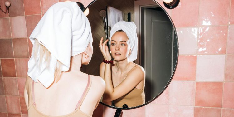 A young woman with a towel in a pink bathroom, reflecting in round mirror as she performs skincare.