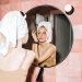 A young woman with a towel in a pink bathroom, reflecting in round mirror as she performs skincare.