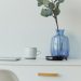 Elegant minimalist desk with blue vase, eucalyptus, mug, and laptop on a white table.