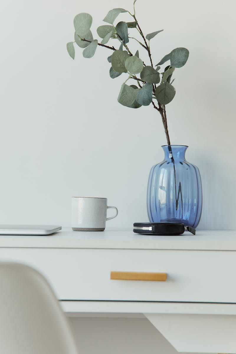 Elegant minimalist desk with blue vase, eucalyptus, mug, and laptop on a white table.