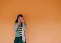 Young woman holding a pink camera stands against an orange wall, exuding a playful fashion vibe.