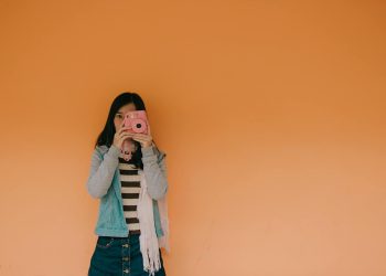 Young woman holding a pink camera stands against an orange wall, exuding a playful fashion vibe.
