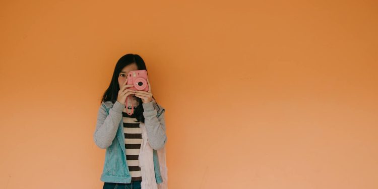Young woman holding a pink camera stands against an orange wall, exuding a playful fashion vibe.
