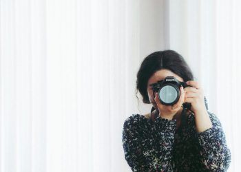 A young woman holding a camera indoors, capturing a photo in a modern setting.