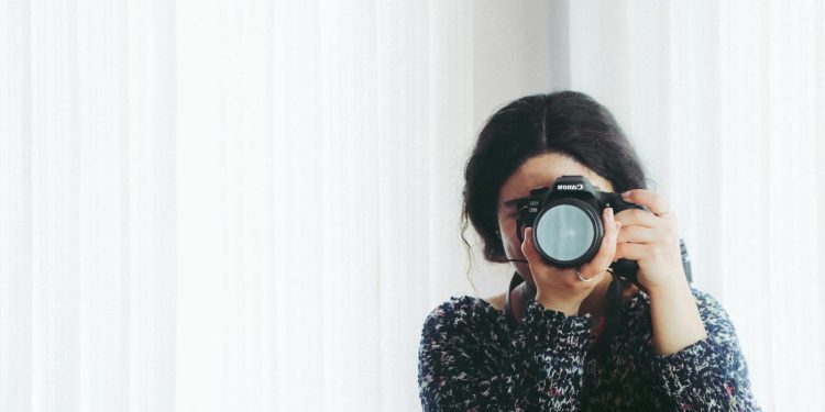 A young woman holding a camera indoors, capturing a photo in a modern setting.