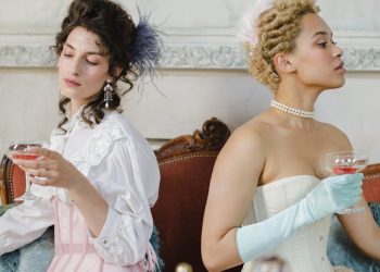 Two elegant women in period costumes enjoying drinks indoors, styled with vintage accessories.