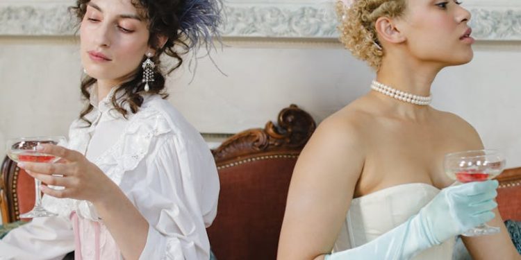 Two elegant women in period costumes enjoying drinks indoors, styled with vintage accessories.