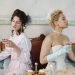 Two elegant women in period costumes enjoying drinks indoors, styled with vintage accessories.